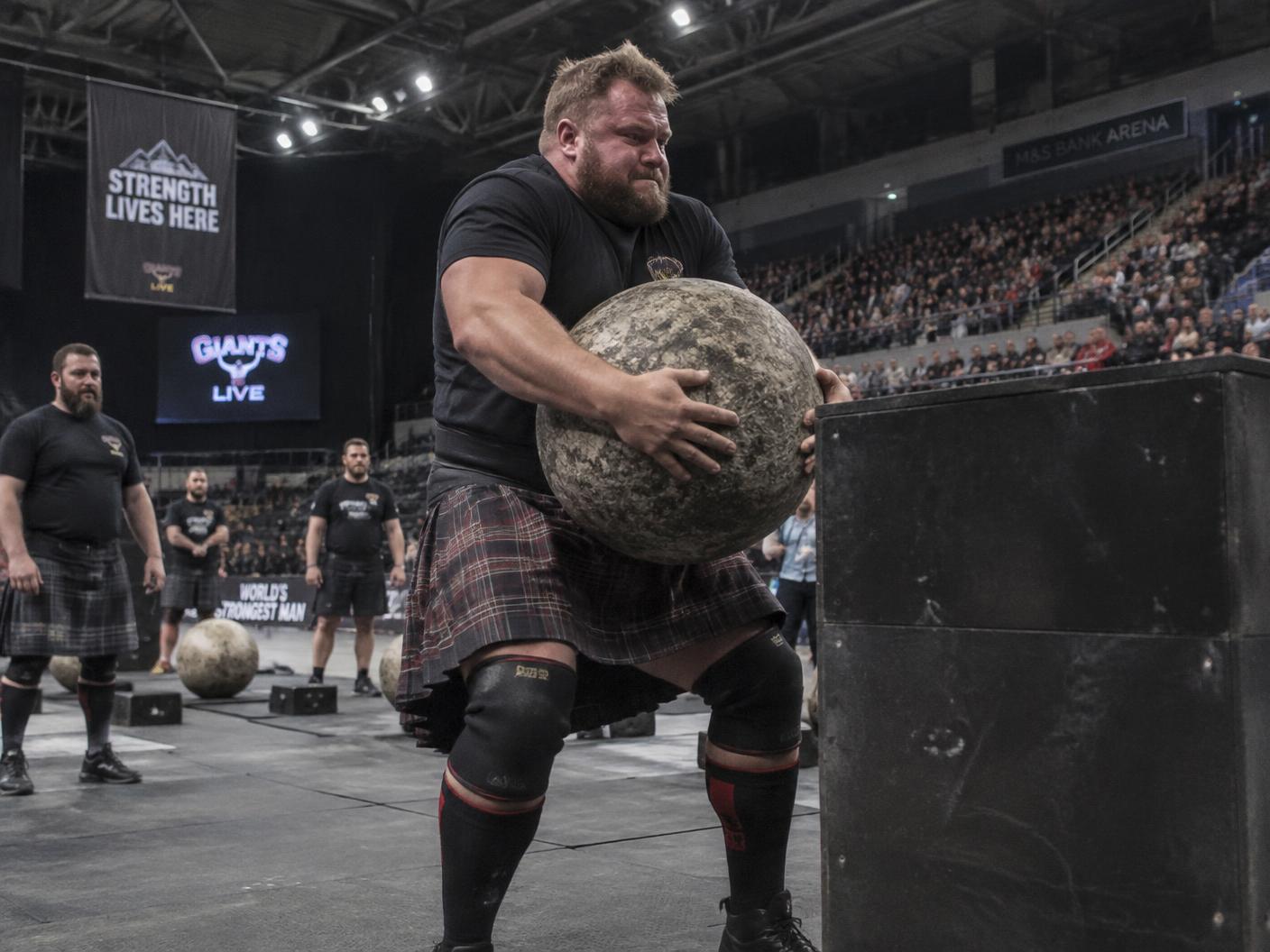 A kilted athlete lifting a heavy stone on an indoor arena floor while a seated crowd watches