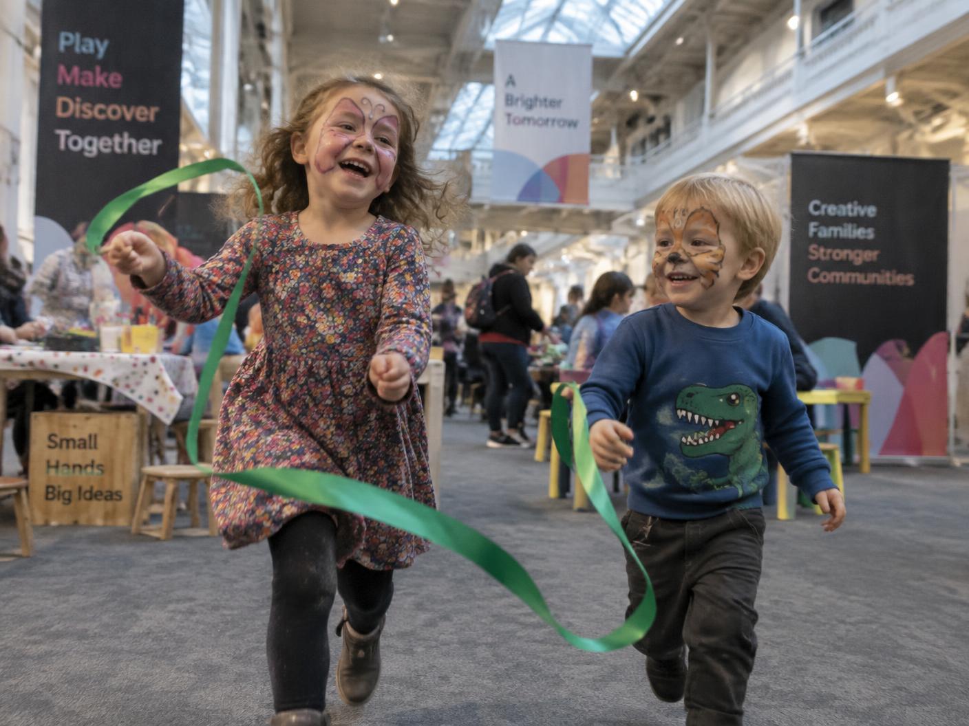 Two small children with painted faces running and laughing with a green ribbon streamer in a bright indoor family area