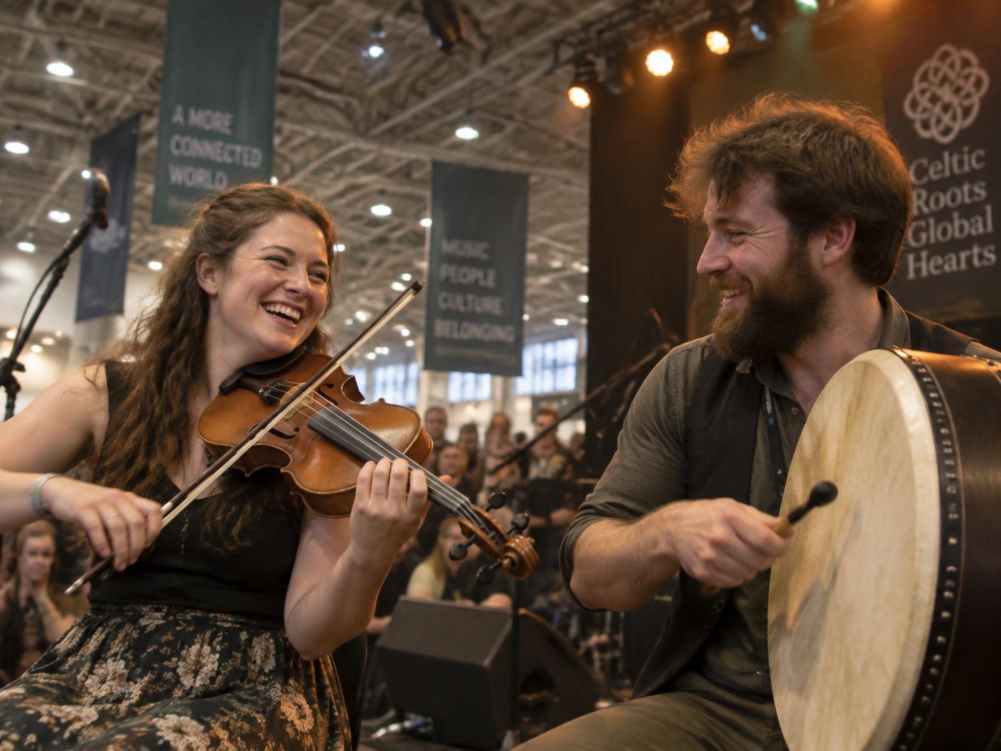A fiddler laughing mid-tune beside a bodhrán player on an indoor hall stage under warm lights